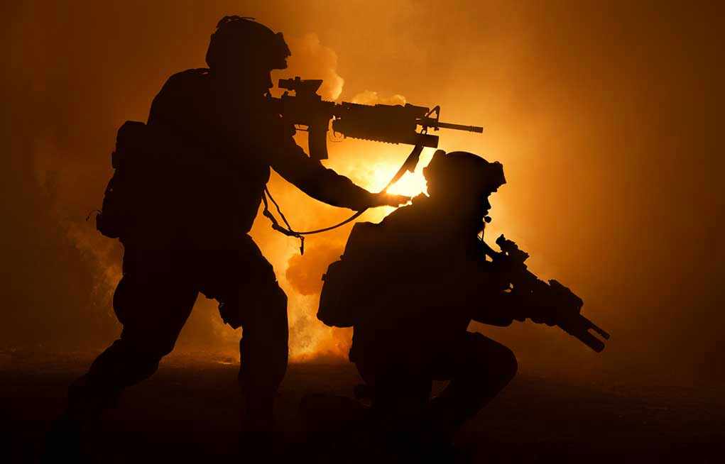 1163416795 Two soldiers with weapons silhouetted against fiery background