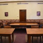 Empty courtroom with wooden tables, chairs, and a door.