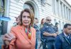 ICE Targets Pelosi – Demand She STANDS DOWN! A woman in an orange blazer speaking passionately at a rally with security personnel in the background