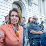 ICE Targets Pelosi – Demand She STANDS DOWN! A woman in an orange blazer speaking passionately at a rally with security personnel in the background