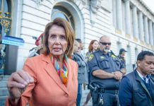 ICE Targets Pelosi – Demand She STANDS DOWN! A woman in an orange blazer speaking passionately at a rally with security personnel in the background