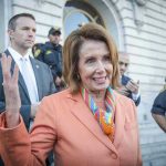 Woman in orange blazer speaking, surrounded by security personnel.