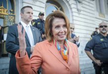 Pelosi’s Mind-Blowing 16,930% – She Made How Much?! Woman in orange blazer speaking, surrounded by security personnel.