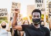BLM Leader INDICTED – 25 Federal Charges Protester raises fist, wears mask, signs in background.