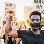 BLM Leader INDICTED – 25 Federal Charges Protester raises fist, wears mask, signs in background.