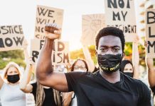 BLM Leader INDICTED – 25 Federal Charges Protester raises fist, wears mask, signs in background.
