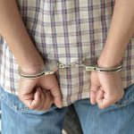 A person in handcuffs viewed from behind, wearing a plaid shirt and denim jeans