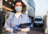 Nurse in scrubs and mask outside hospital holding clipboard.