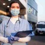 Nurse in scrubs and mask outside hospital holding clipboard.