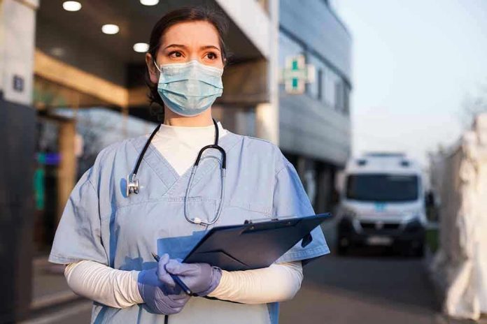 1714681855 Nurse in scrubs and mask outside hospital holding clipboard.
