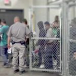 Violence Erupts – Feds CLASH With Anti-ICE Protestors! People standing inside chain-link holding area with an officer nearby