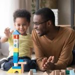 Father and child building a tall tower with colorful wooden blocks on the floor
