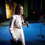 Dems Beg AOC to ‘Stay Away’ in Swing-States Woman in a light suit holding a microphone on stage