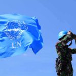 UN peacekeeper with blue helmet looks through binoculars beside UN flag