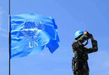 UN peacekeeper with blue helmet looks through binoculars beside UN flag