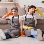 Two tired cleaners sitting on the floor by a sofa
