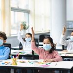 Schoolchildren wearing masks raise hands in a classroom