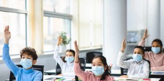 Schoolchildren wearing masks raise hands in a classroom
