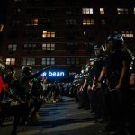 Achille Emaná kneels with raised fist facing a line of police officers at night