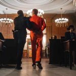 Prisoner in orange jumpsuit escorted by officer in a courtroom