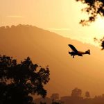 Tourist Flight CRASHES Into Mountains – 13 KILLED! Small airplane silhouetted at sunset over tree-lined hills