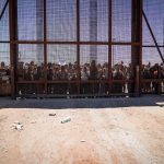 U.S. Issues Urgent Travel Warning – Avoid Region Crowd stands behind tall metal fence at border crossing