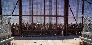 Crowd stands behind tall metal fence at border crossing