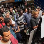 Crowd gathers around a woman reading a notice board at a bus stop