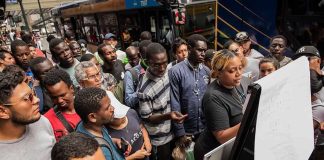 Crowd gathers around a woman reading a notice board at a bus stop
