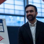 Mamdani Appoints Controversial Child Youtube Star To TOP Job Zohran Mamdani smiling indoors near a voting sign with an American flag