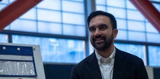 Zohran Mamdani smiling indoors near a voting sign with an American flag