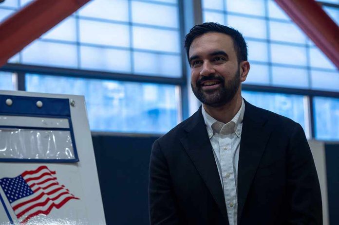 Zohran Mamdani smiling indoors near a voting sign with an American flag