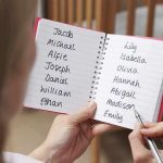 Hands holding a notebook with baby name lists near a crib
