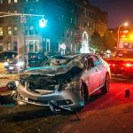 Wrecked silver car on city street at night with tow truck nearby
