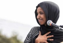Ilhan Omar speaking at a microphone with hand on chest