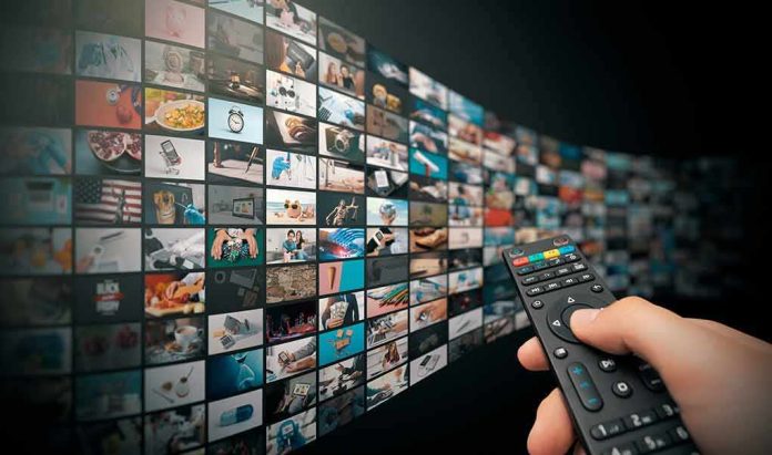 shutterstock_1921373024 Hand holding remote control facing a wall of streaming thumbnails