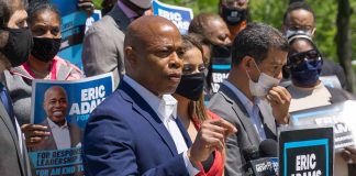 Man speaking at campaign event with supporters behind him.