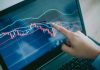 Hand pointing at stock market graph on laptop.