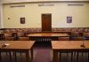 Empty courtroom with wooden tables, chairs, and a door.