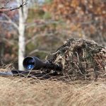 A sniper rifle partially covered in camouflage netting in a grassy area